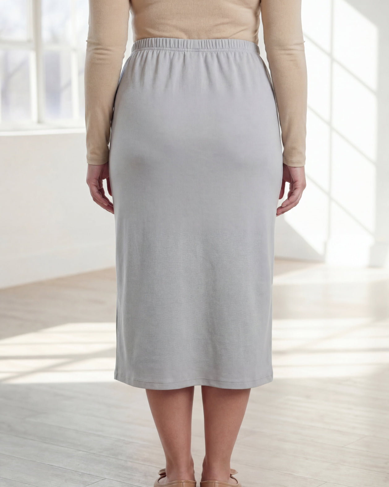 Woman wearing a beige top and silver skirt in a bright room with large windows.