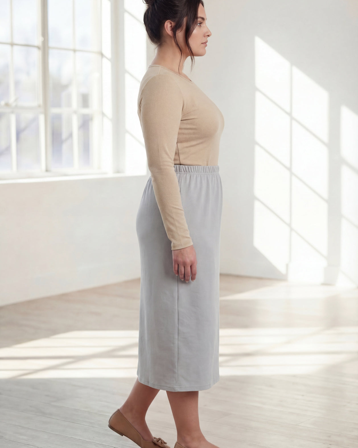 Woman wearing a beige top and silver skirt in a bright room with large windows.