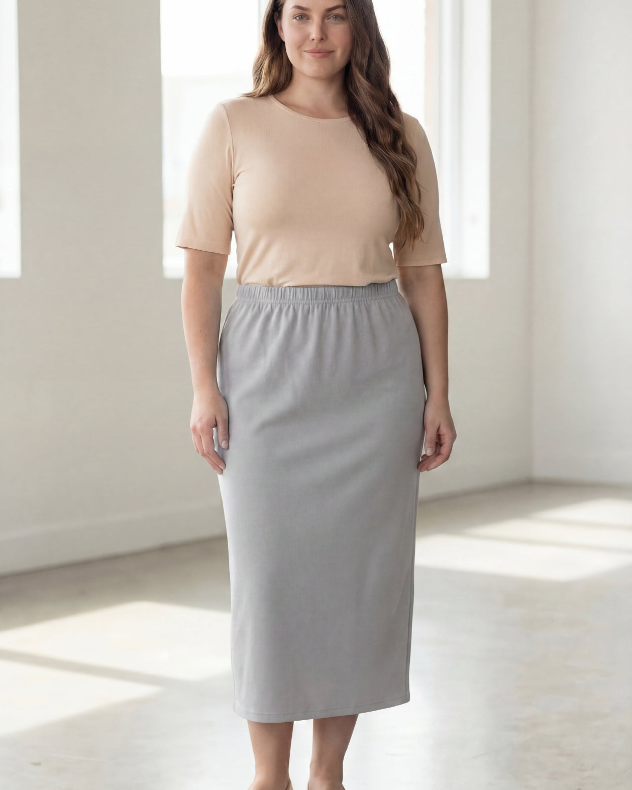 Woman wearing a beige shirt & silver maxi length pencil skirt standing in a bright room with large windows.