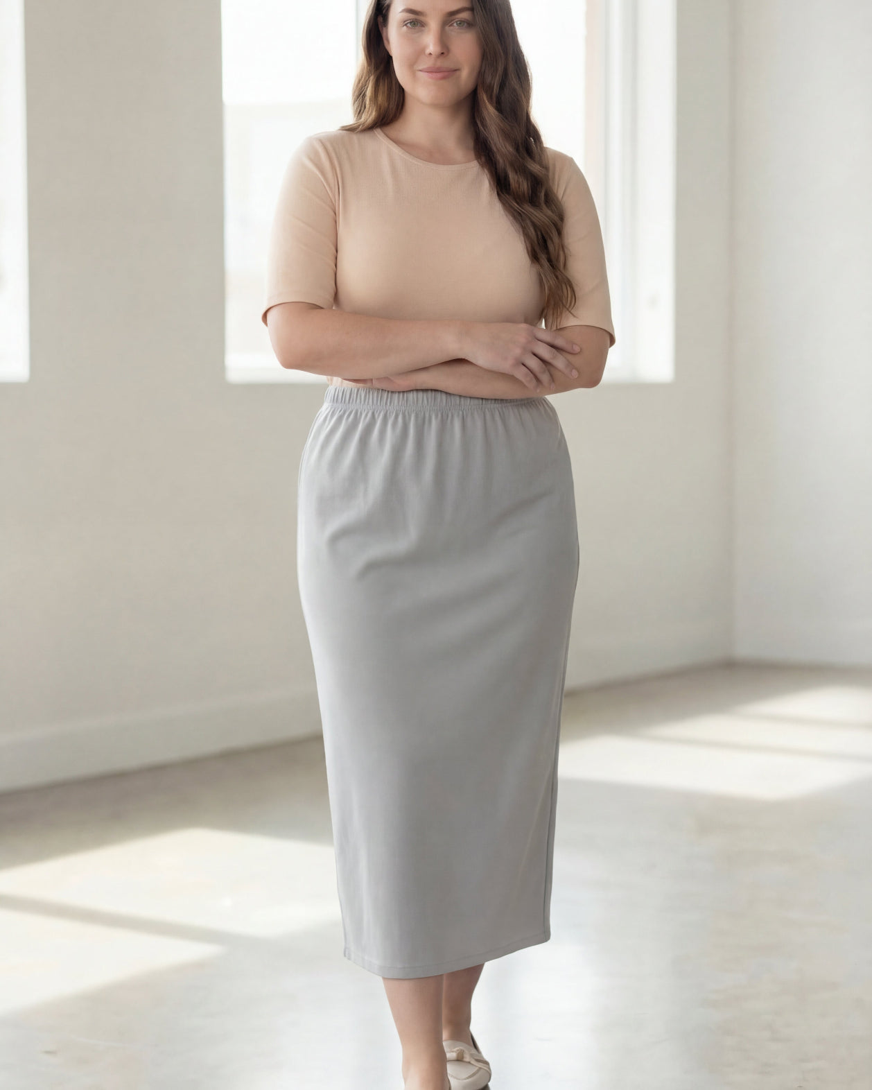 Woman wearing a beige shirt & silver maxi length pencil skirt standing in a bright room with large windows.