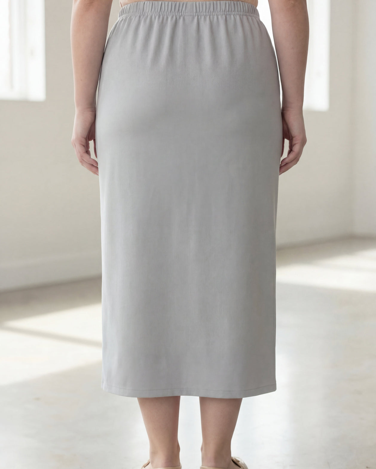 Woman wearing a beige shirt & silver maxi length pencil skirt standing in a bright room with large windows.