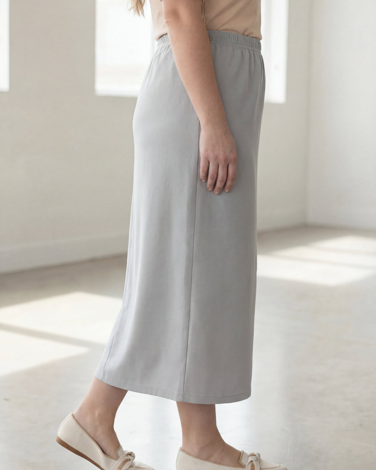 Woman wearing a beige shirt & silver maxi length pencil skirt standing in a bright room with large windows.