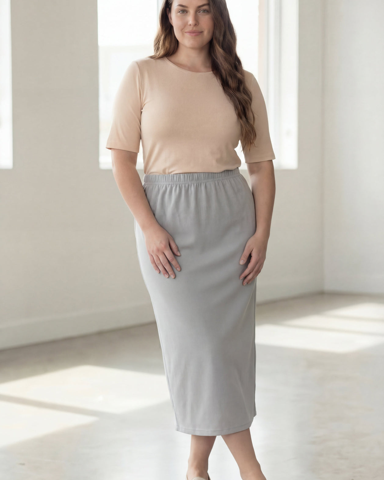 Woman wearing a beige shirt & silver maxi length pencil skirt standing in a bright room with large windows.