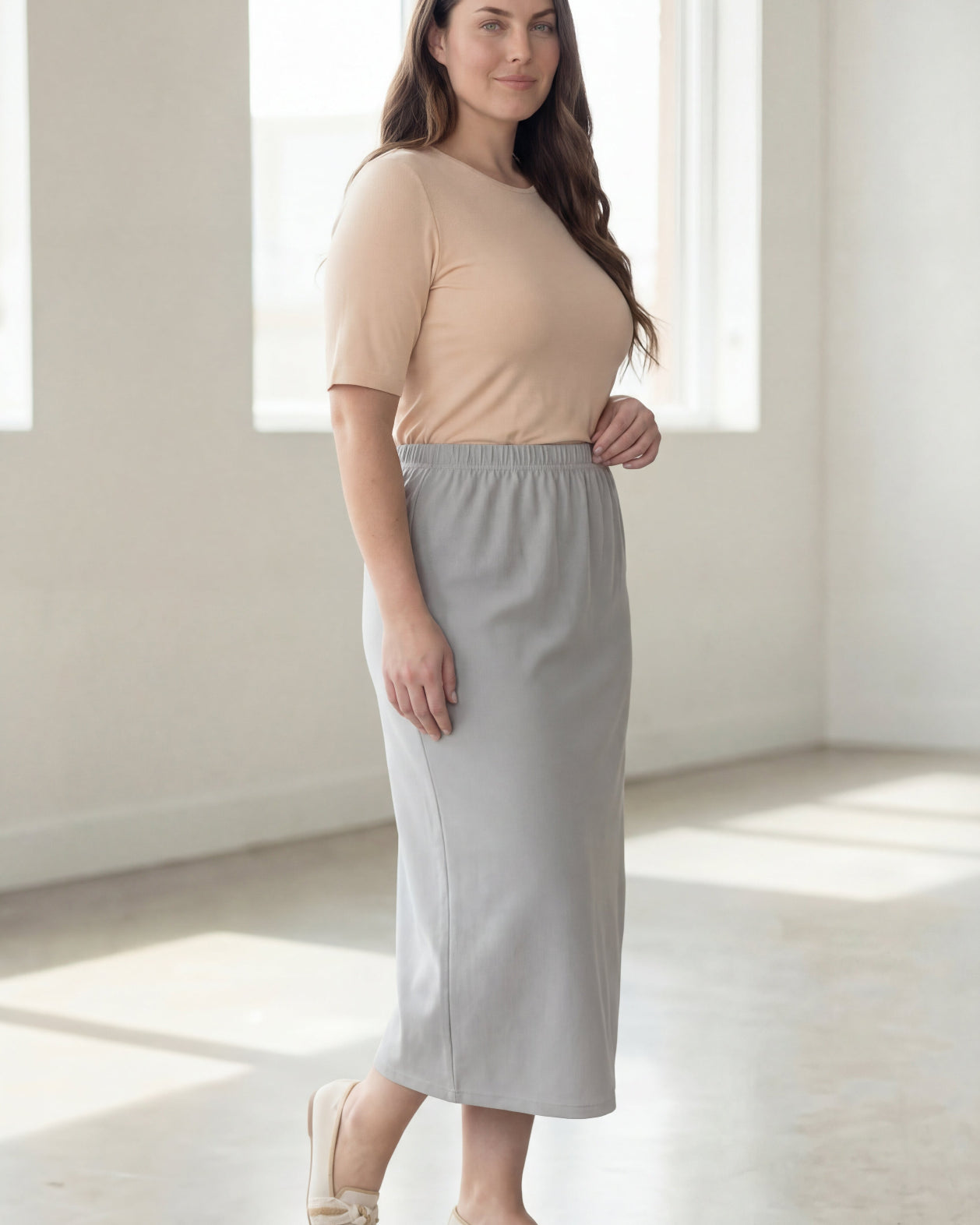 Woman wearing a beige shirt & silver maxi length pencil skirt standing in a bright room with large windows.