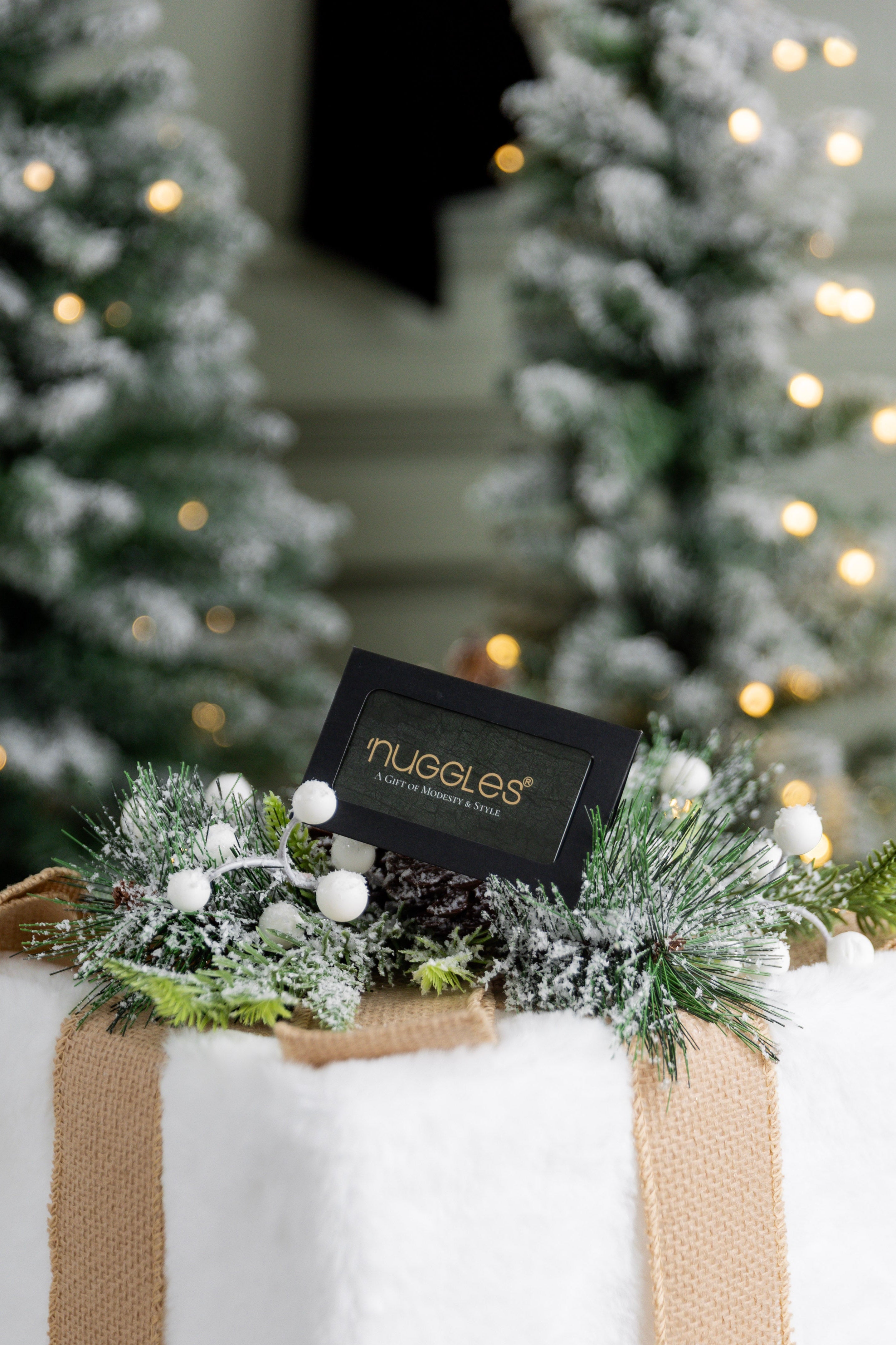 Gift box wrapped in white paper and burlap ribbon with a small Nuggles® giftcard, surrounded by Christmas trees and lights.