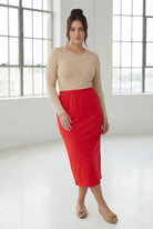 Woman wearing a beige top and red skirt in a bright room with large windows.