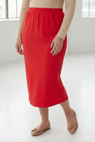 Woman wearing a beige top and red skirt in a bright room with large windows.