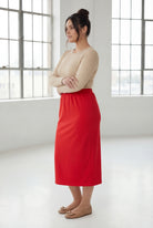 Woman wearing a beige top and red skirt in a bright room with large windows.