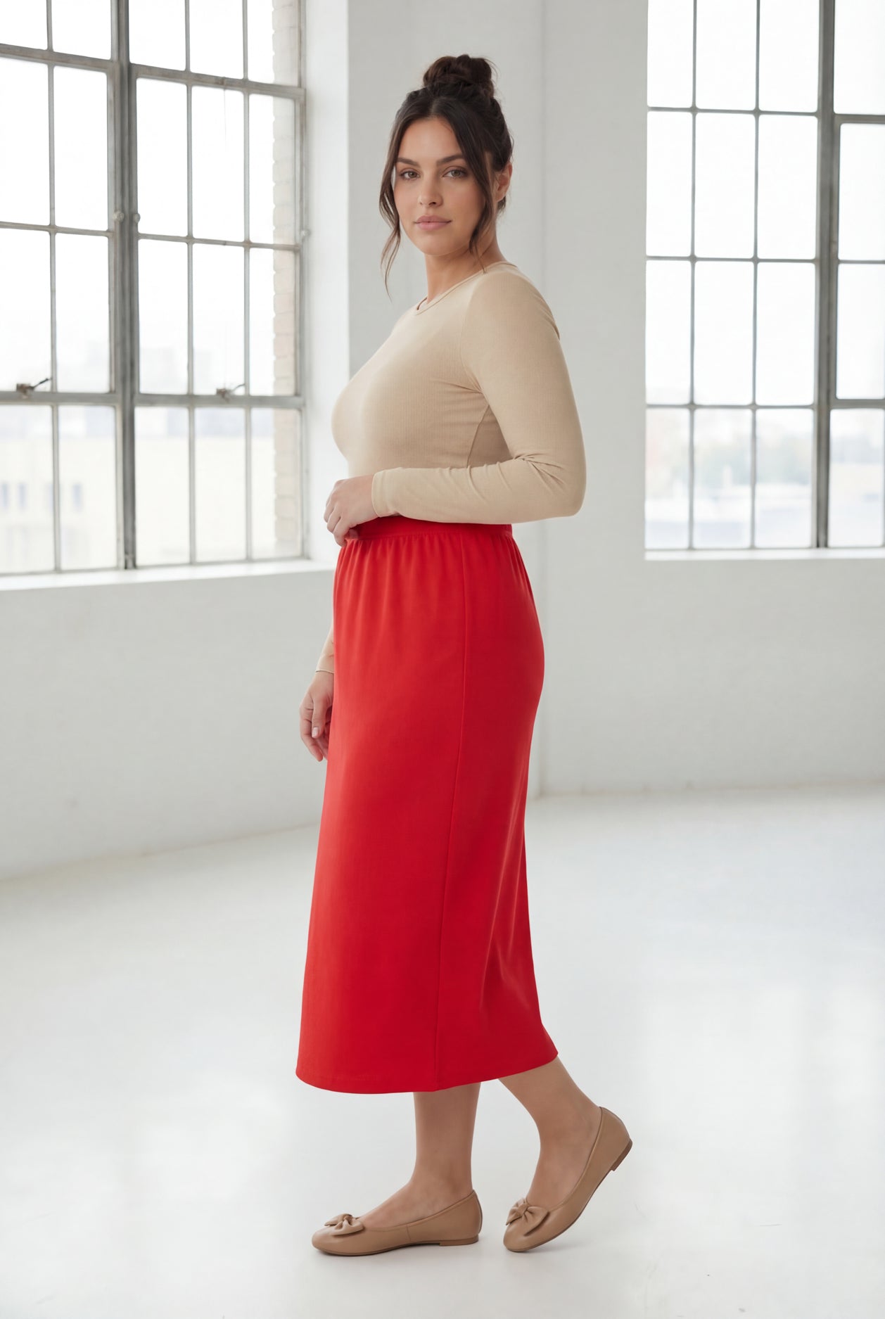 Woman wearing a beige top and red skirt in a bright room with large windows.