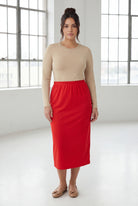 Woman wearing a beige top and red skirt in a bright room with large windows.