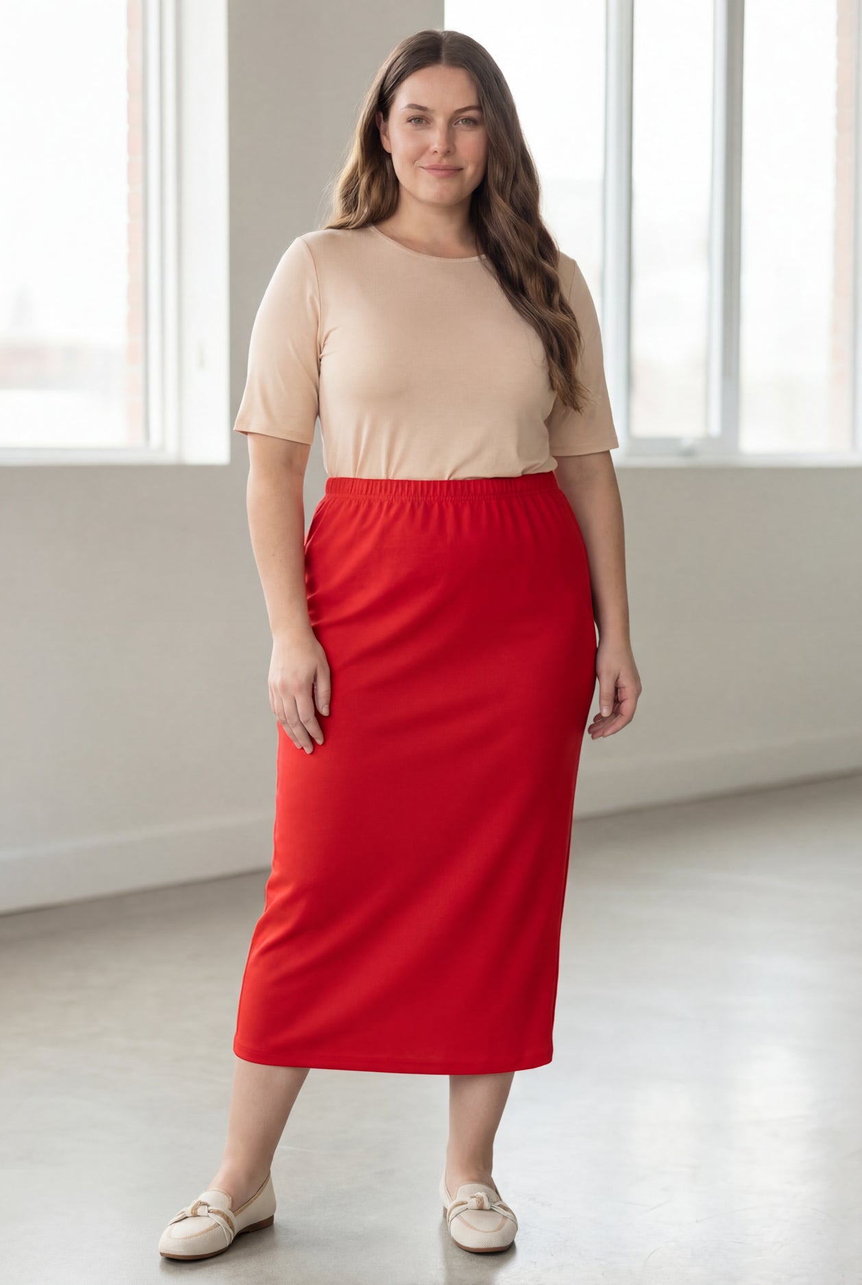 Woman wearing a beige shirt & red maxi length pencil skirt standing in a bright room with large windows.