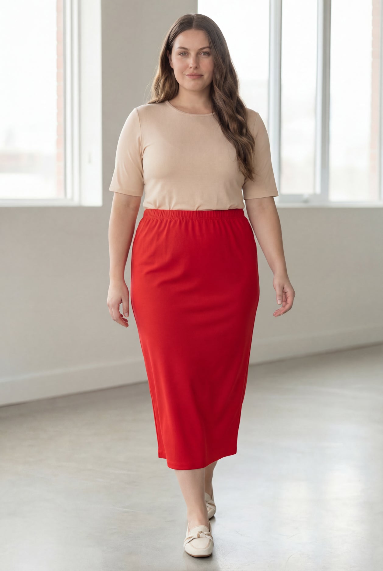 Woman wearing a beige shirt & red maxi length pencil skirt standing in a bright room with large windows.