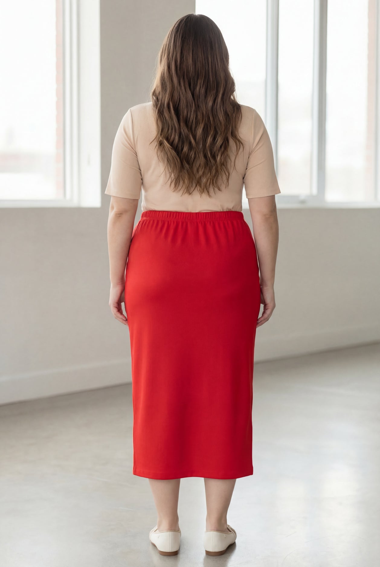 Woman wearing a beige shirt & red maxi length pencil skirt standing in a bright room with large windows.