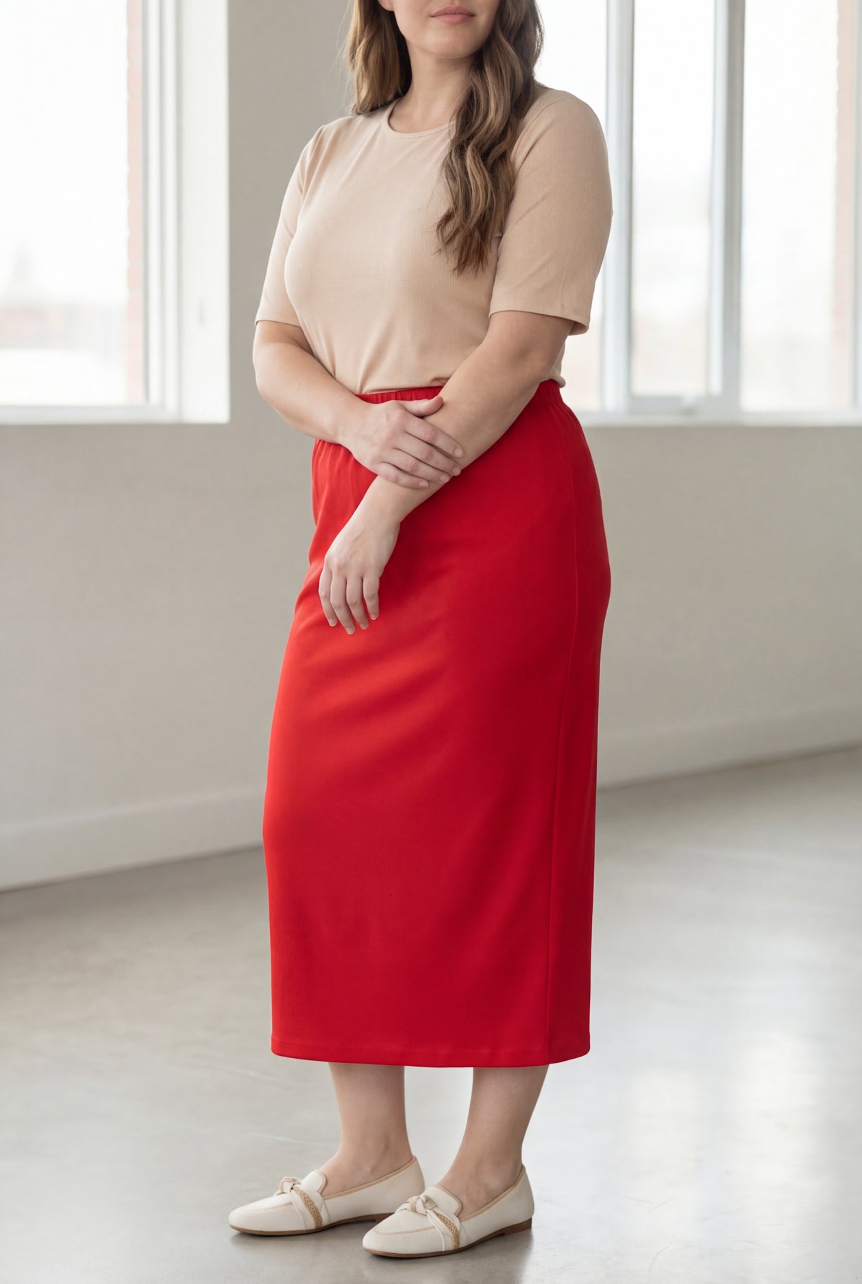 Woman wearing a beige shirt & red maxi length pencil skirt standing in a bright room with large windows.