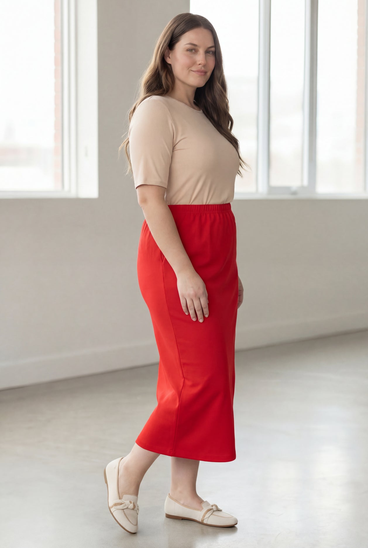 Woman wearing a beige shirt & red maxi length pencil skirt standing in a bright room with large windows.