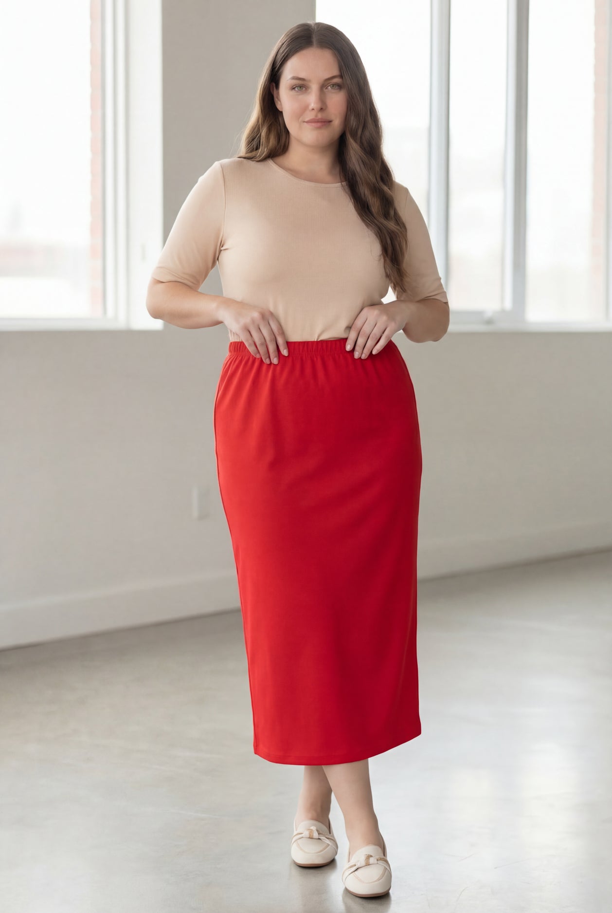 Woman wearing a beige shirt & red maxi length pencil skirt standing in a bright room with large windows.
