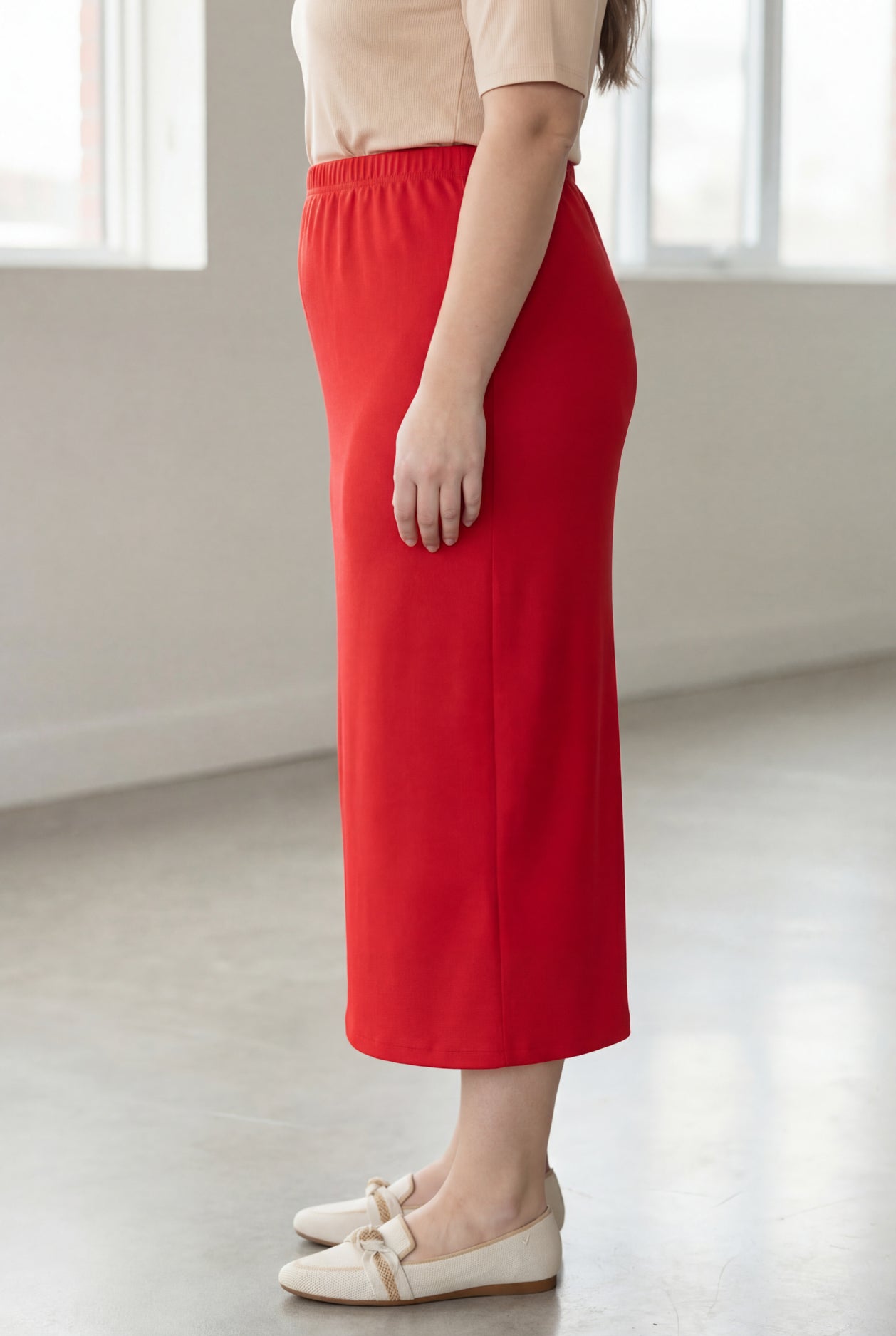 Woman wearing a beige shirt & red maxi length pencil skirt standing in a bright room with large windows.
