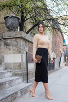 Woman in beige top and black skirt standing on a city street.
