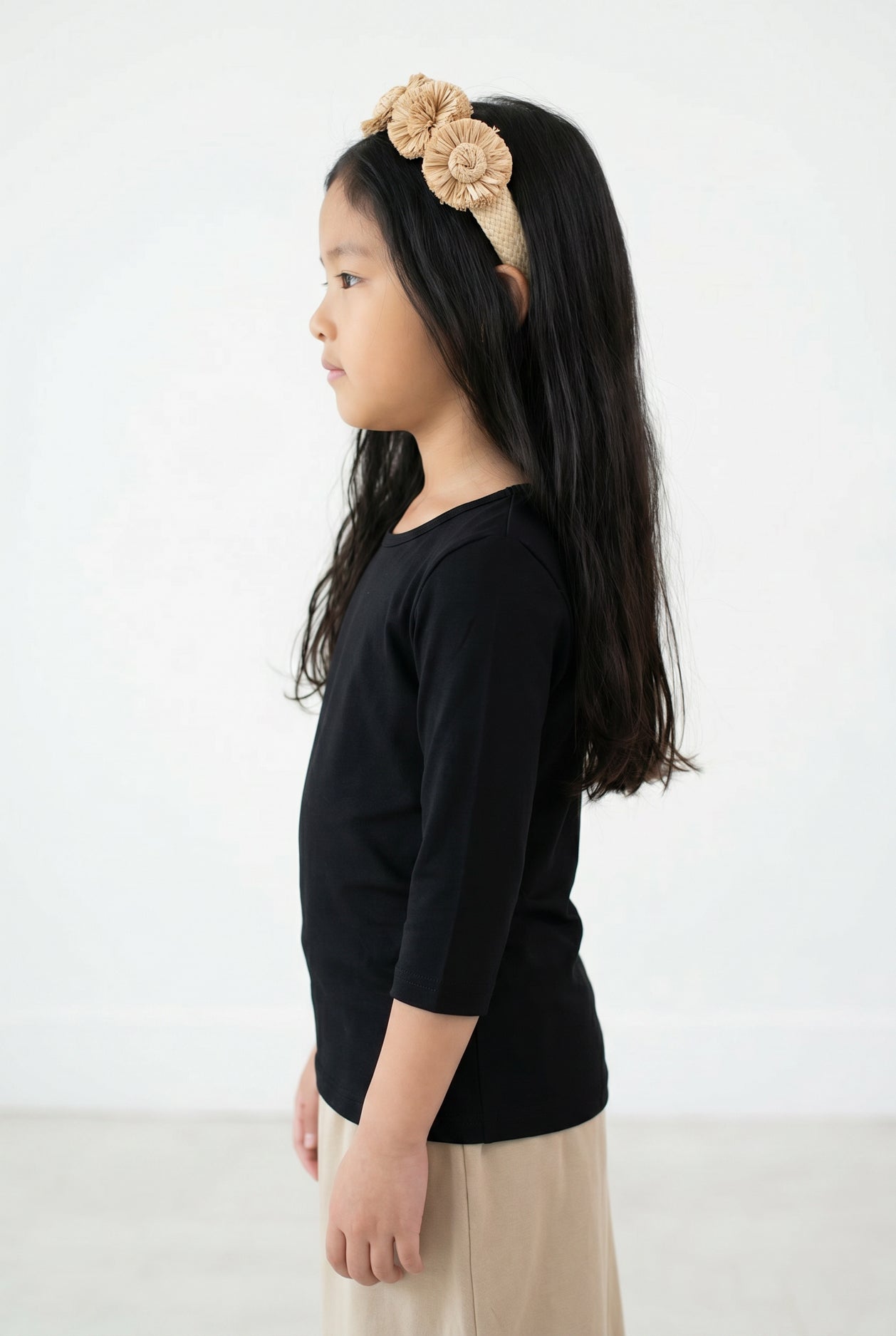 Young girl with long dark hair wearing Nuggles® Black Tee and a beige floral headband against a white background. 