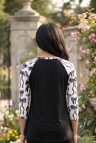 Person wearing a black and white patterned top standing in a garden with flowers and stone pillars.