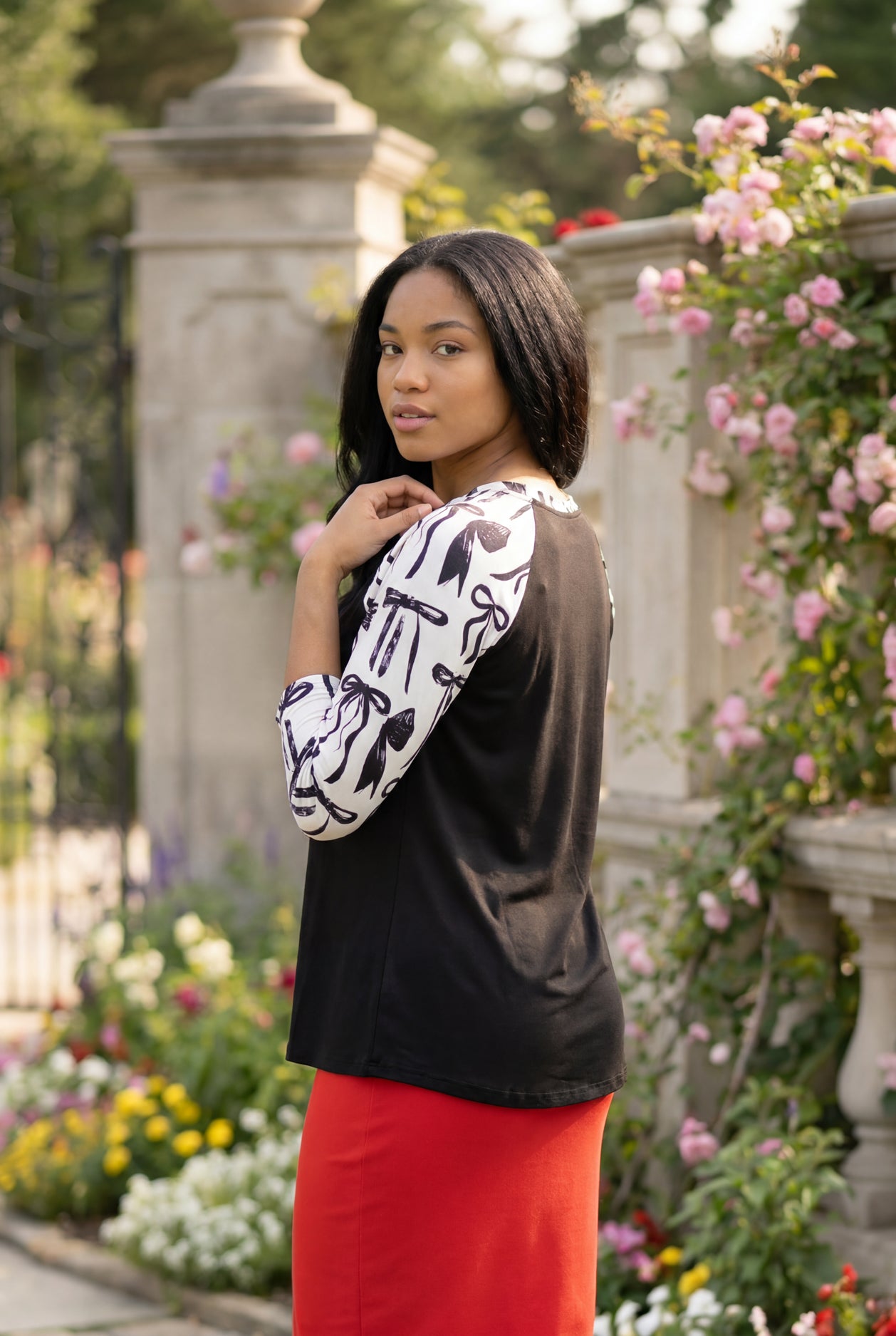 Person wearing a black and white patterned top standing in a garden with flowers and stone pillars.