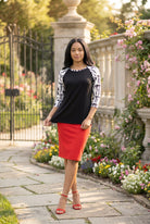 Person wearing a black and white patterned top standing in a garden with flowers and stone pillars.