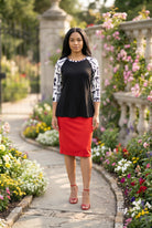 Person wearing a black and white patterned top standing in a garden with flowers and stone pillars.