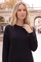 Nuggles® Oakley Hoodie Dress in Black, waist-up view with a hand at the ribbed crew neckline in a stone courtyard