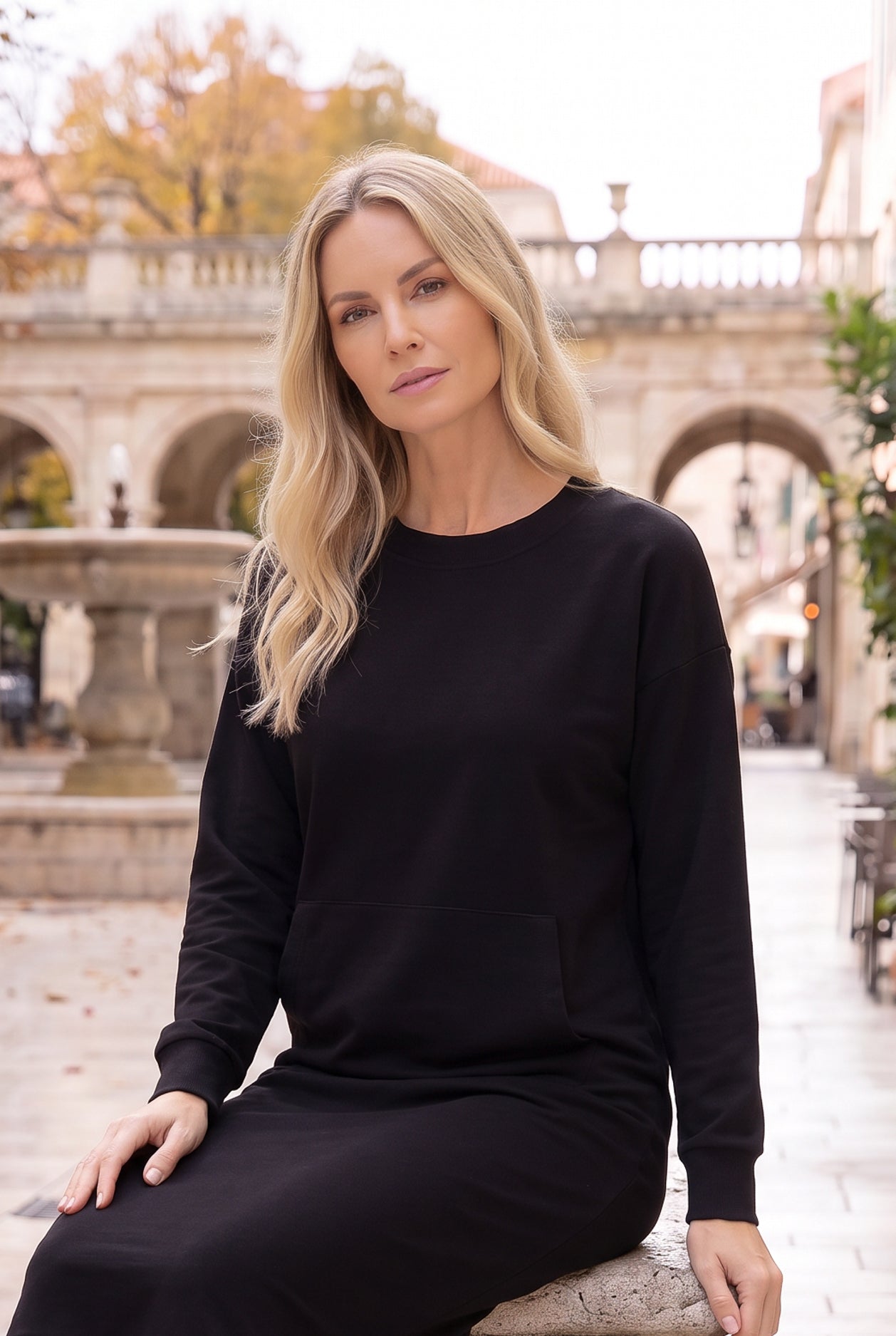 Nuggles® Oakley Hoodie Dress in Black, seated on a stone ledge beside a courtyard fountain