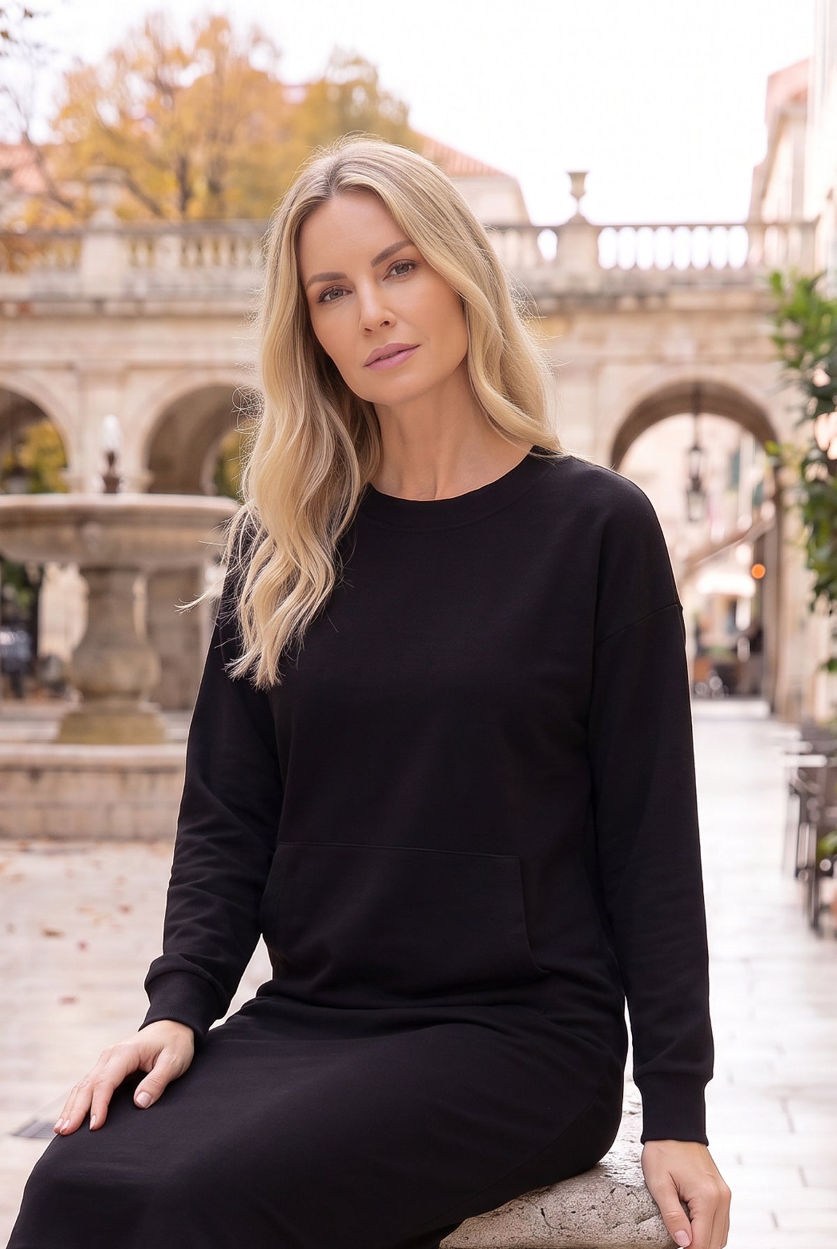 Nuggles® Oakley Hoodie Dress in Black, seated on a stone ledge beside a courtyard fountain