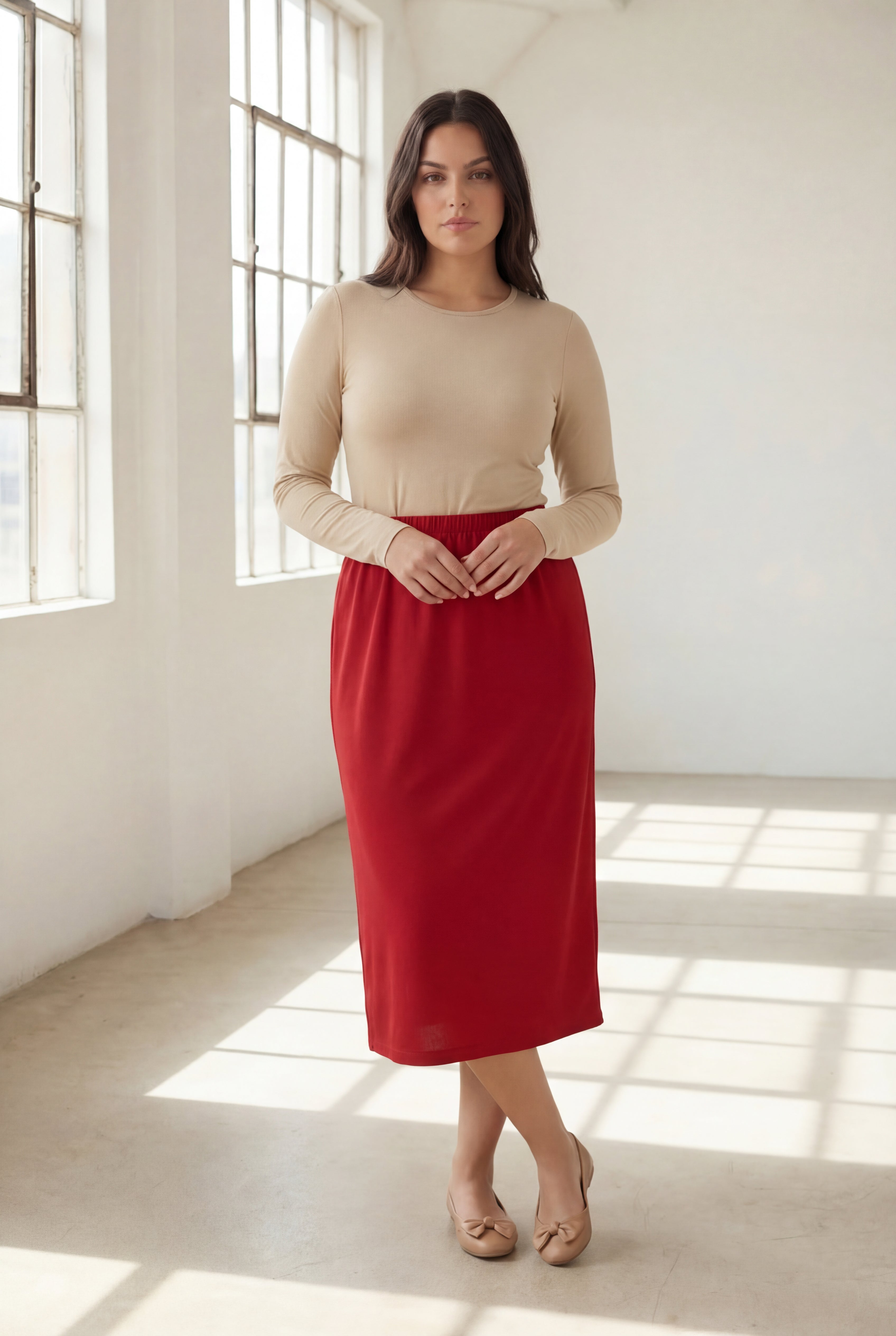 Woman wearing a beige top and red skirt standing in a bright room with large windows.