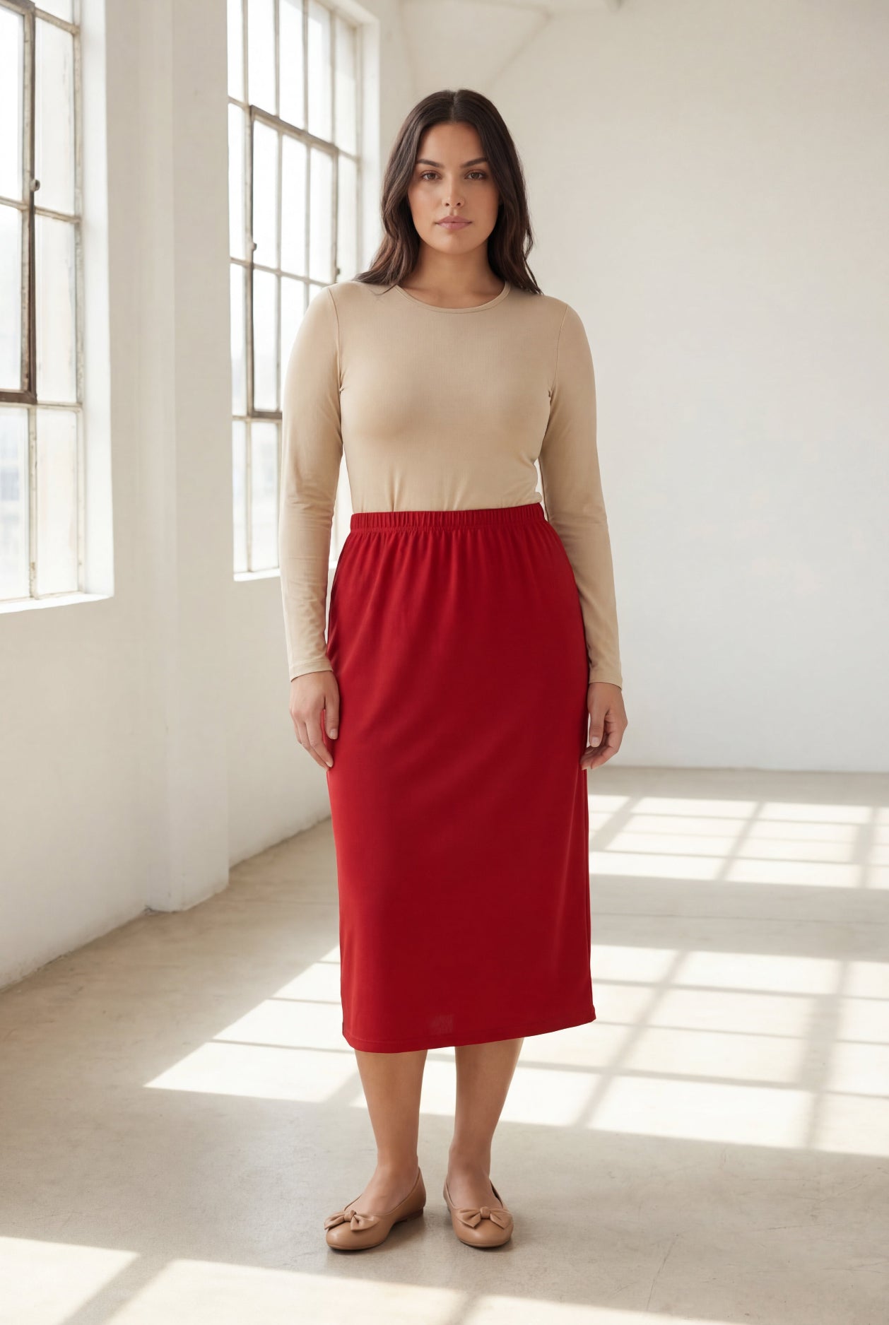 Woman wearing a beige top and red skirt standing in a bright room with large windows.