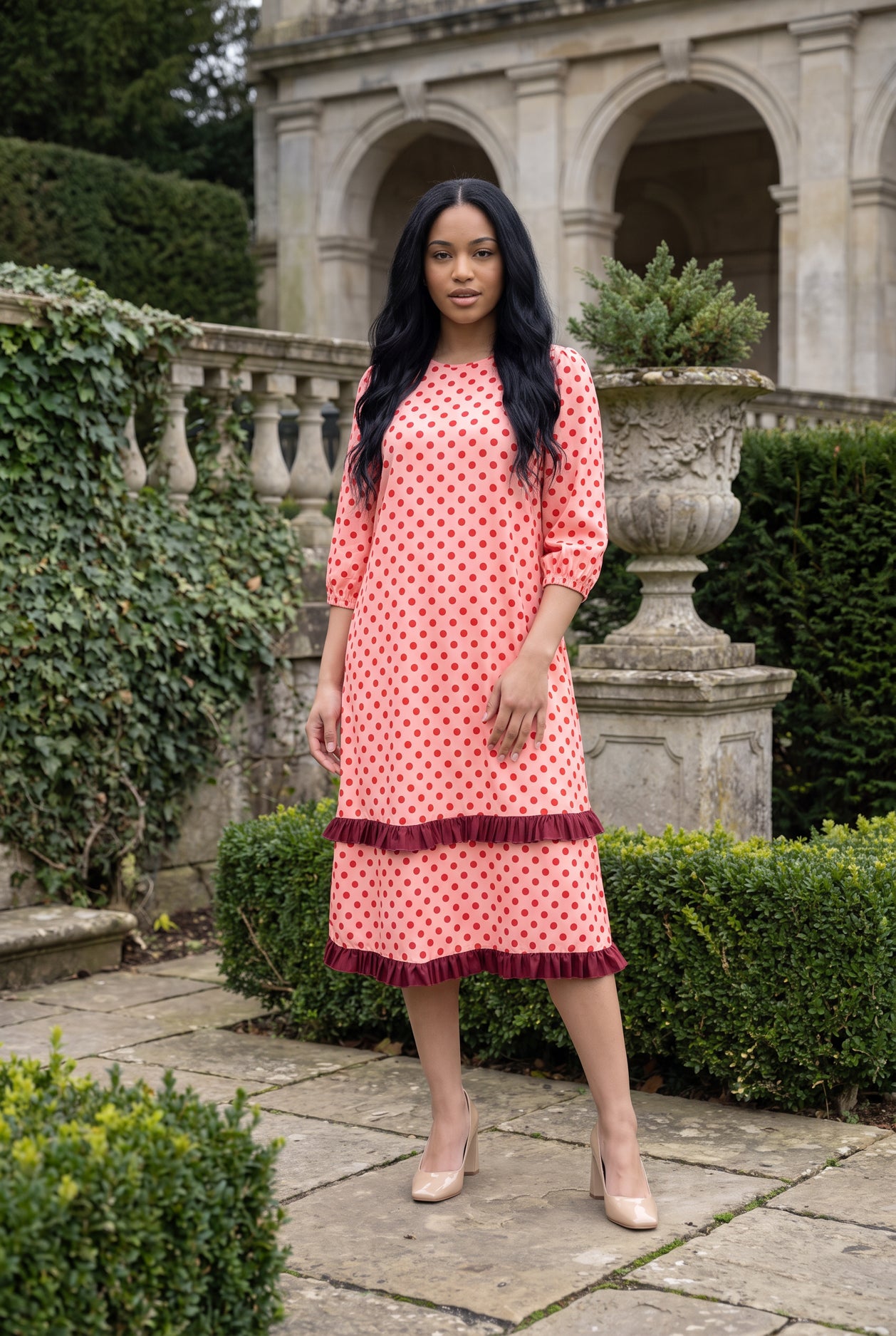 Nuggles® Patricia Dress in Cherry Dot, full length on a garden terrace, red dots on a coral ground