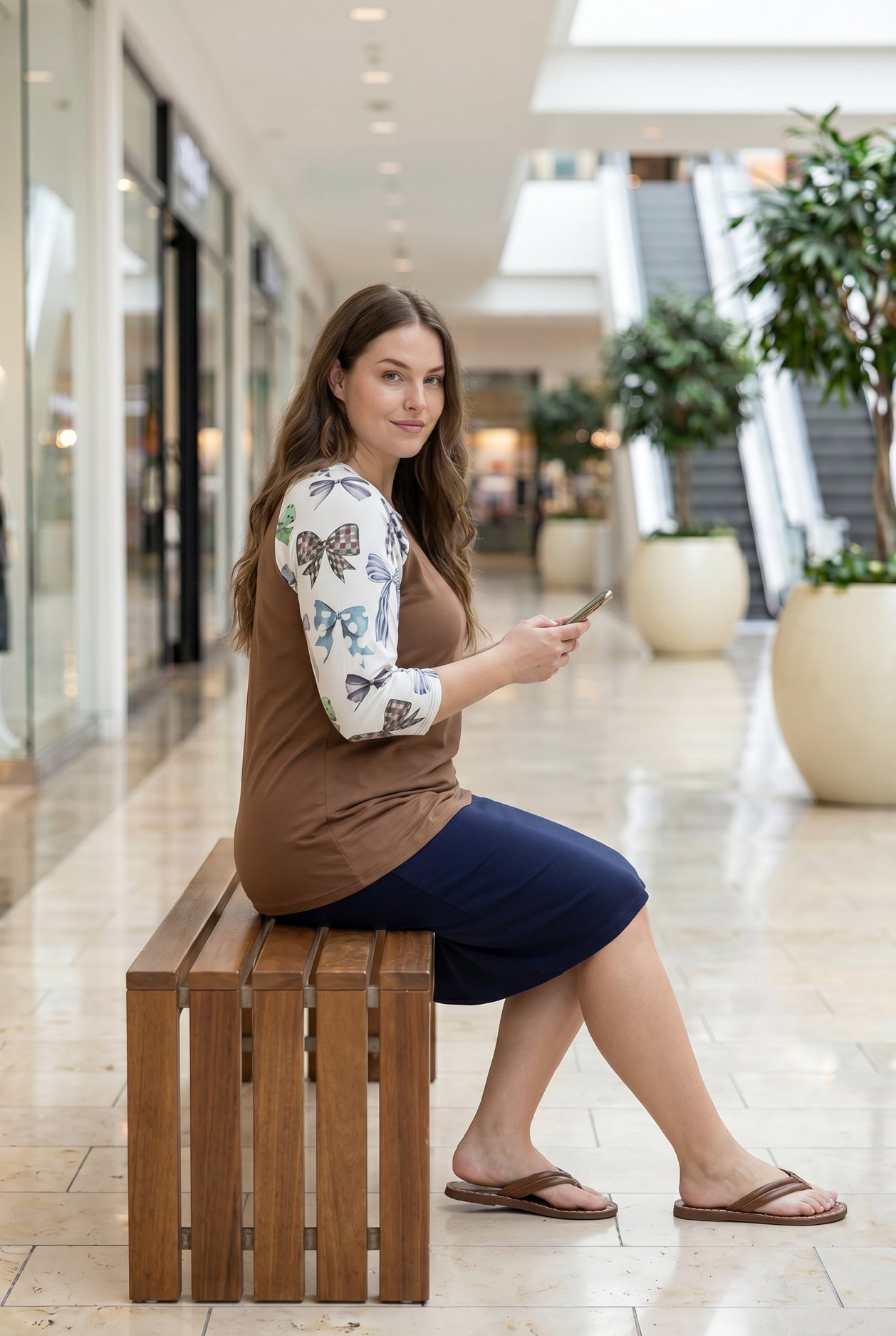 Woman modeling a chestnut brown top with white ribbon-print sleeves and a navy skirt in a bright indoor shopping mall setting.