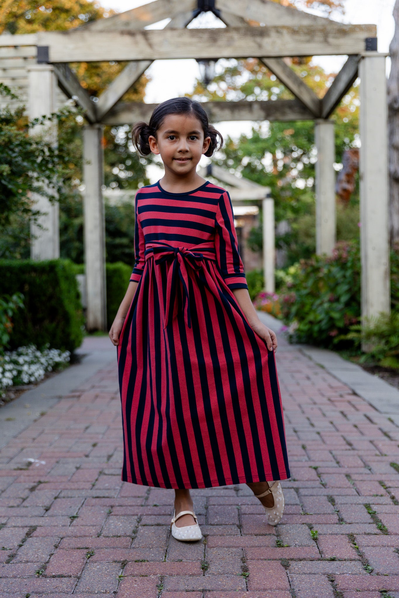 Young girl wearing the Nuggles® Girls Adelia Dress — a modest striped maxi dress with bow tie waist.