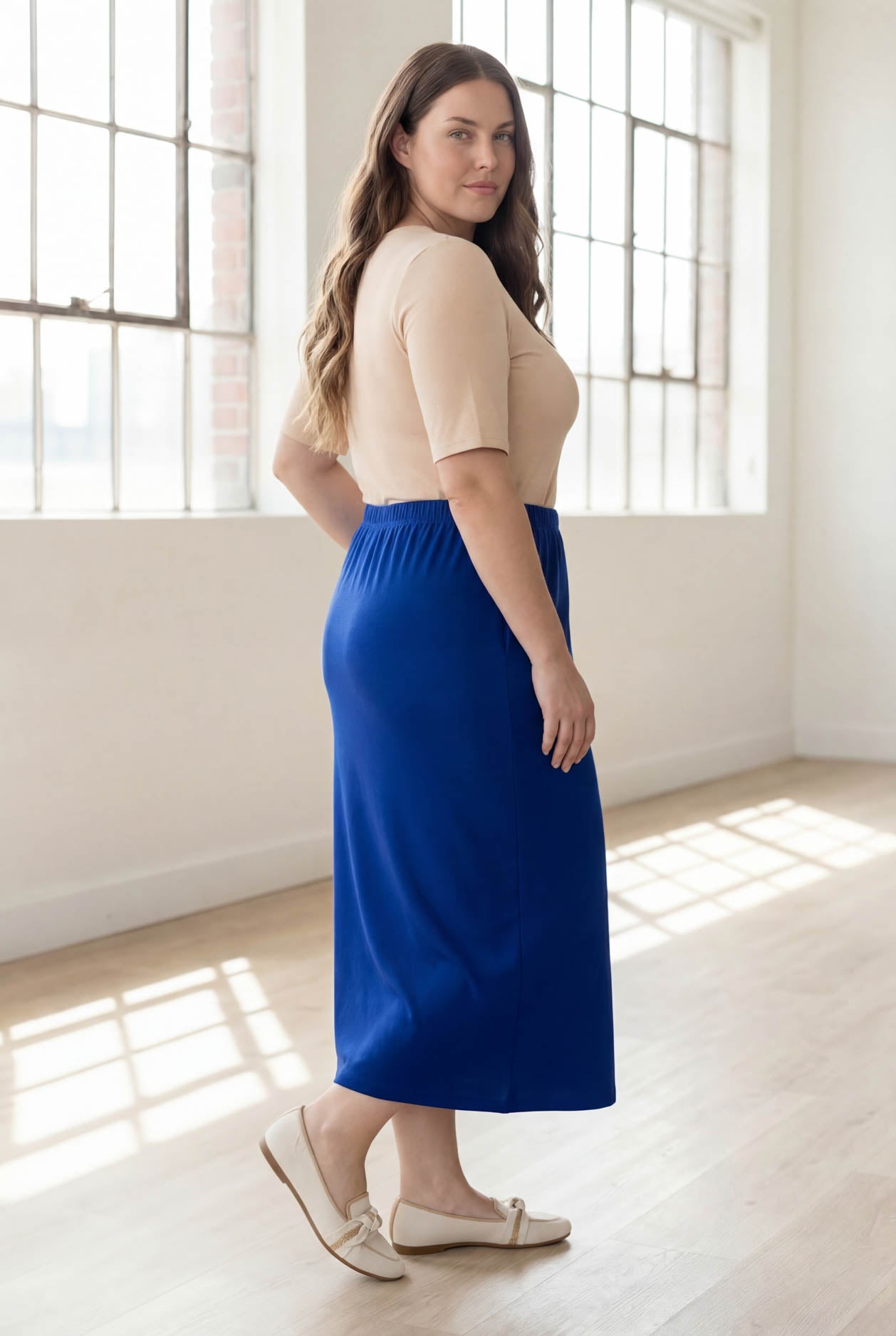 Woman wearing a blue skirt and beige top in a bright room with large windows.