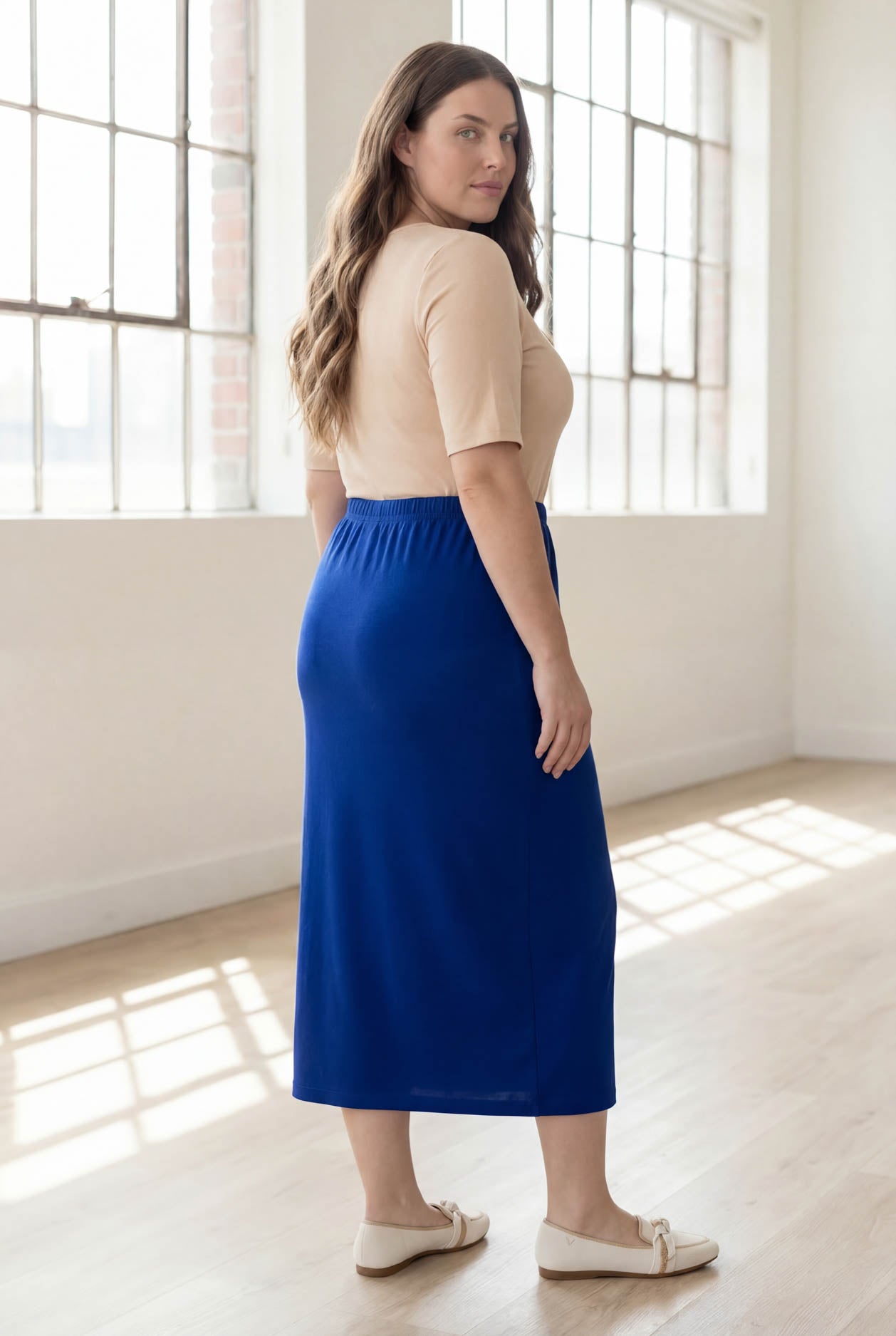 Woman wearing a blue skirt and beige top in a bright room with large windows.