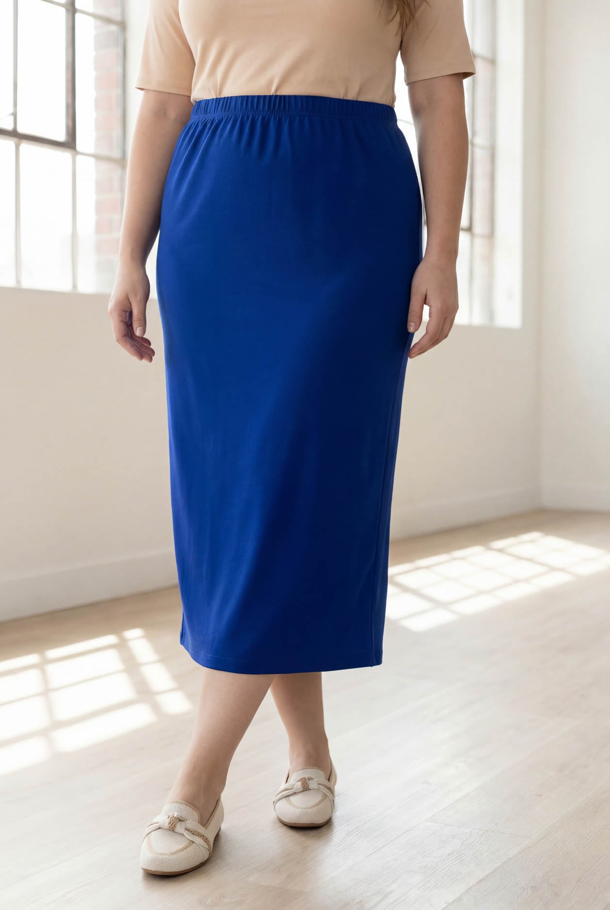 Person wearing a blue skirt and beige top in a bright room with large windows.