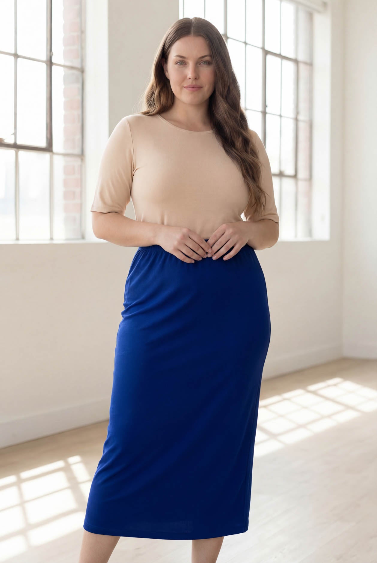 Woman wearing a beige top and blue skirt standing in a bright room with large windows.