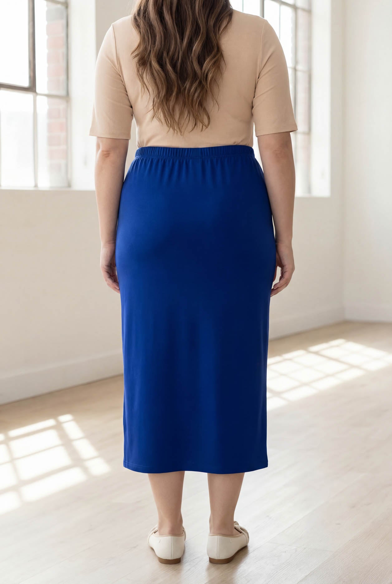 Person wearing a blue skirt and beige top in a bright room with large windows.