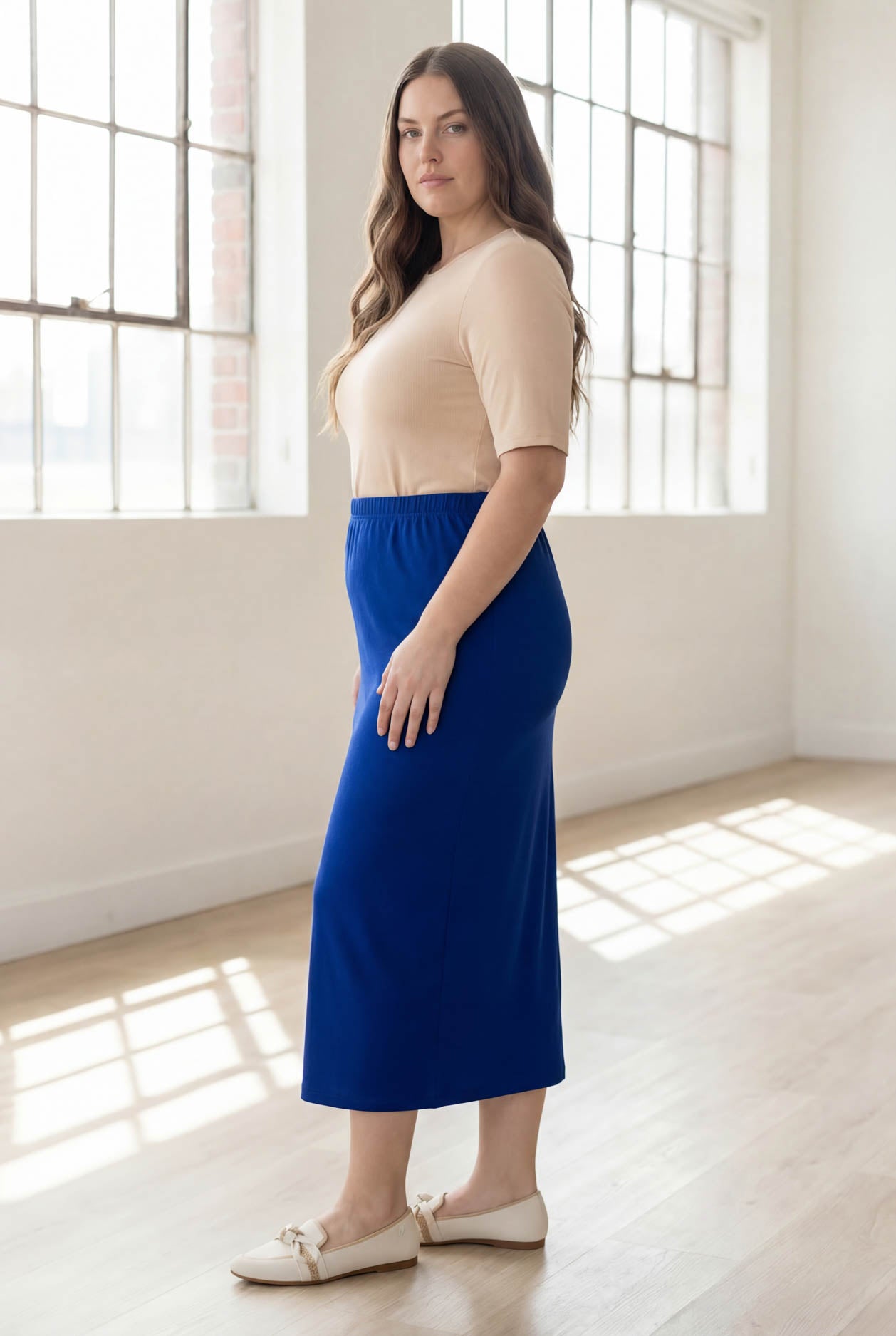 Woman wearing a blue skirt and beige top in a bright room with large windows.