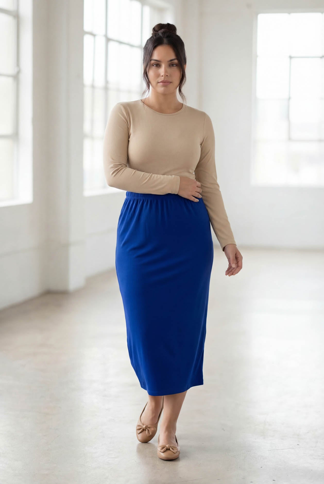 Woman wearing a beige long-sleeve top and blue skirt in a bright room.