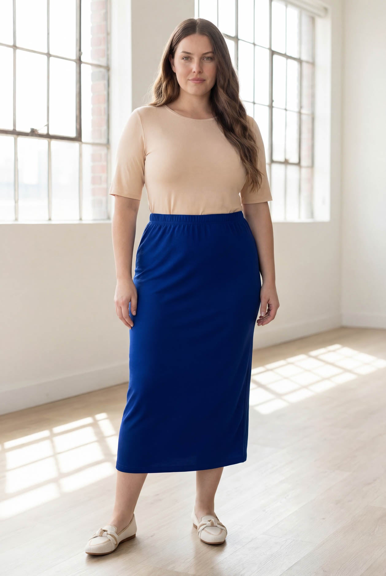 Woman wearing a beige top and blue skirt in a bright room with large windows.