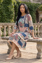 Nuggles® Patricia Dress in English Garden, seated full length with the tiered skirt draped over a stone bench