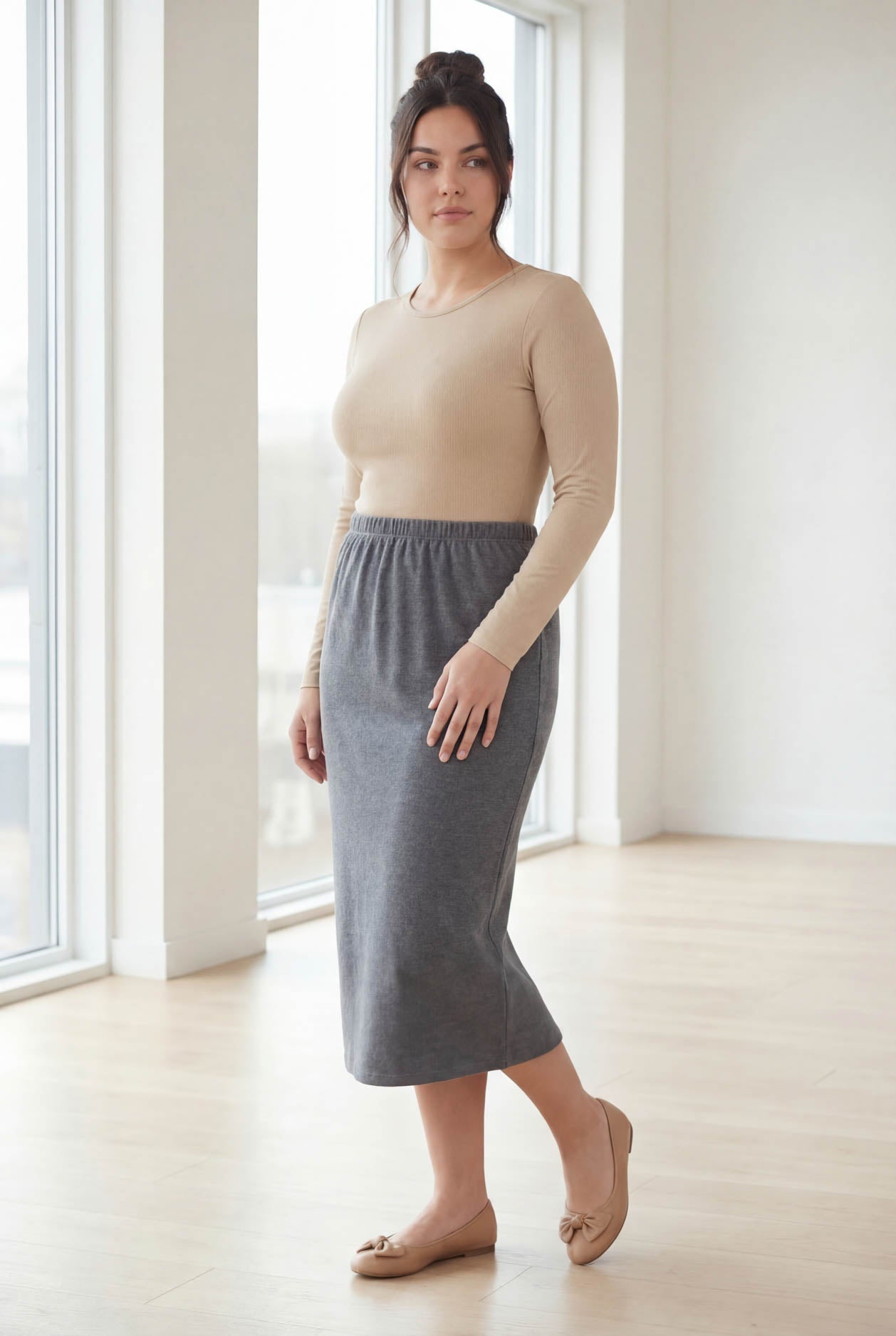 Woman wearing a beige top and gray skirt standing in a bright room with large windows.