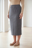 Woman wearing a beige top and gray skirt standing in a bright room with large windows.