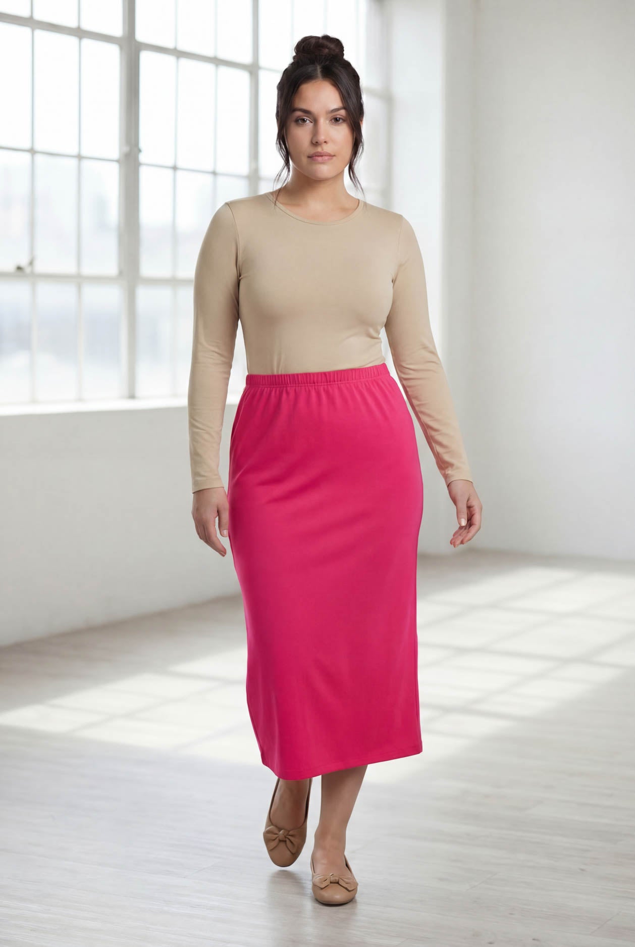 Woman wearing a beige long-sleeve top and pink skirt in a bright room.