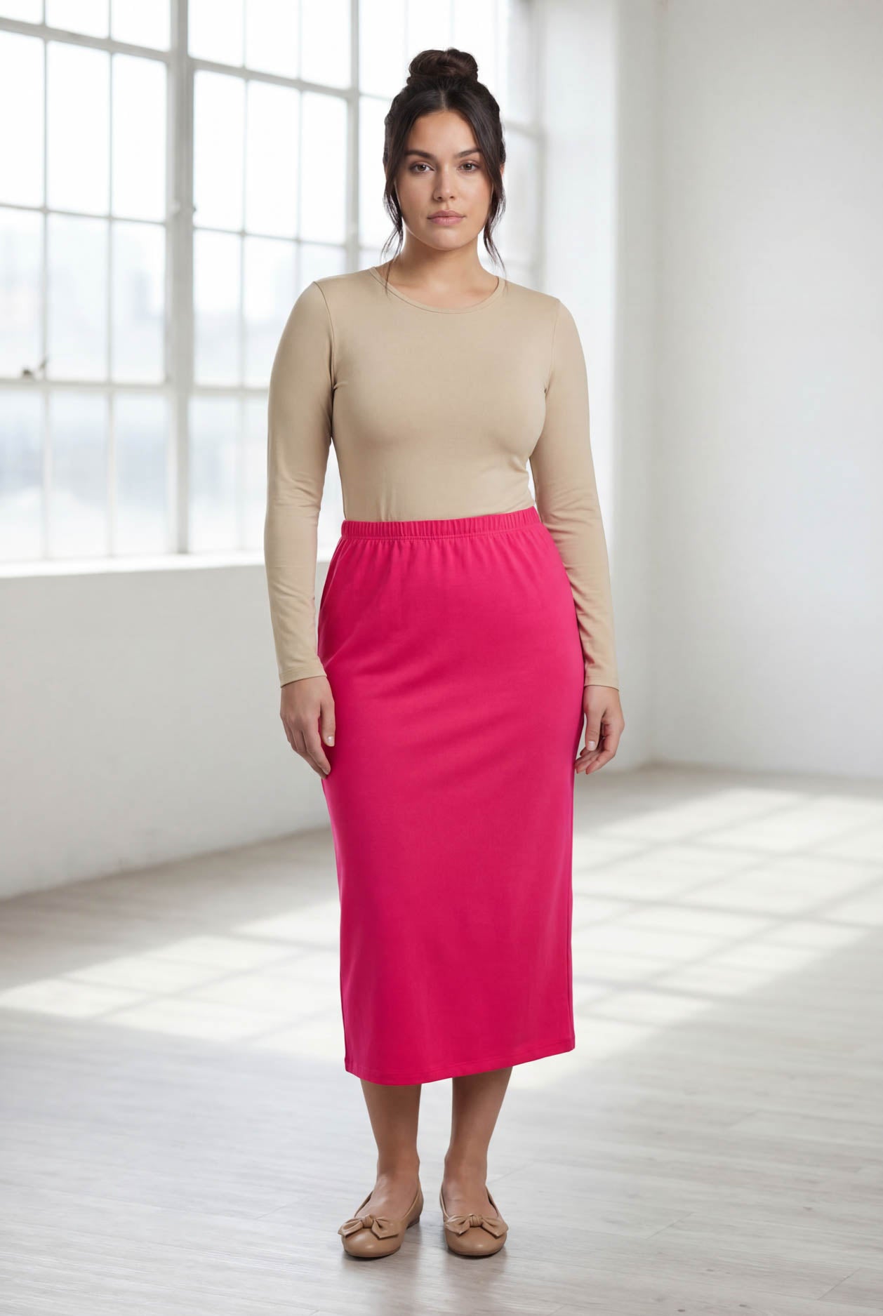 Woman wearing a beige long-sleeve top and pink skirt in a bright room.