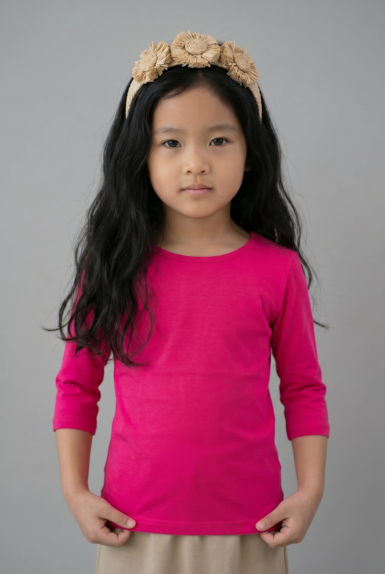 Young girl wearing a bright pink long-sleeve shirt and beige headband with flowers on a gray background