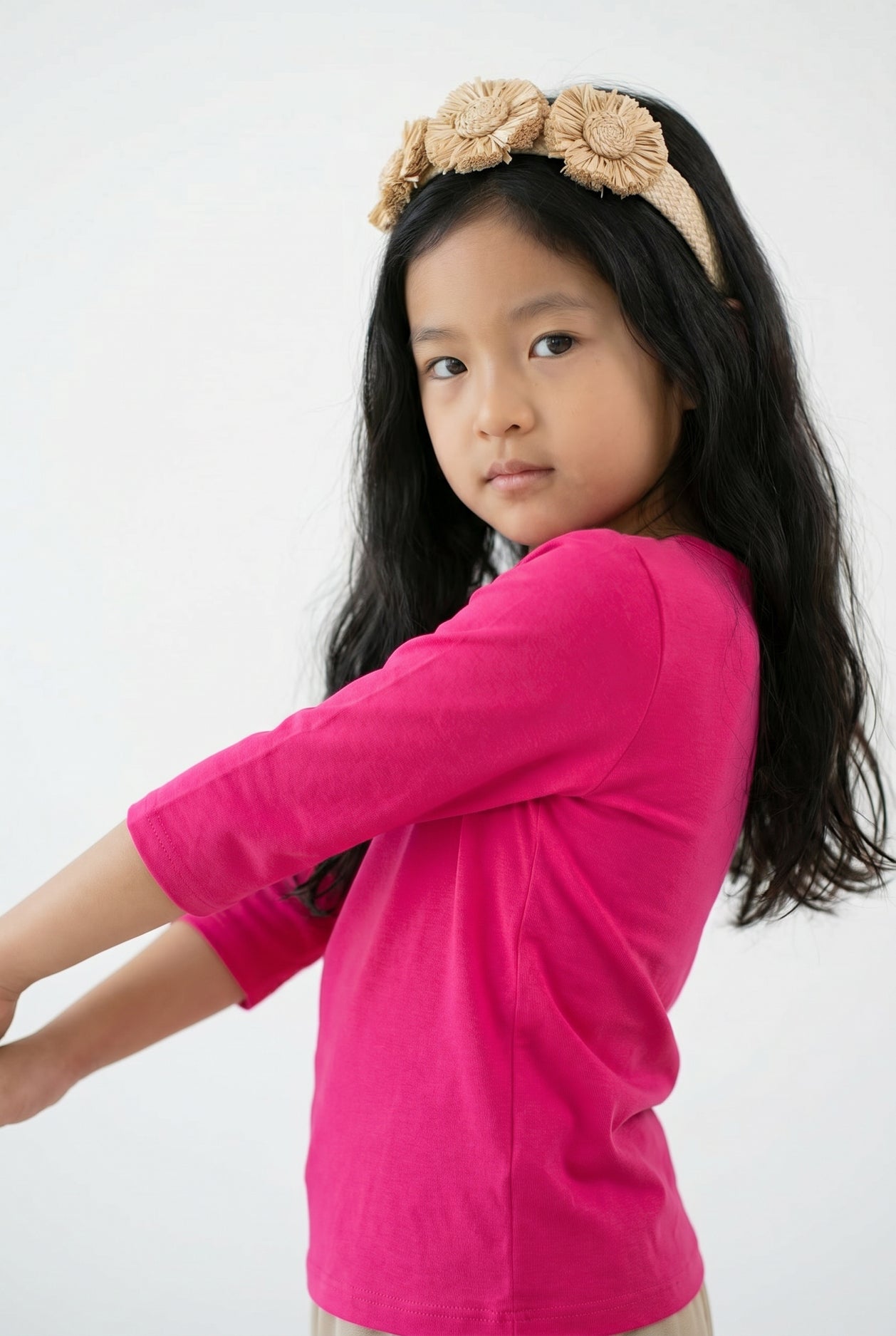 Young girl wearing a bright pink long-sleeve shirt and beige headband with flowers on a gray background
